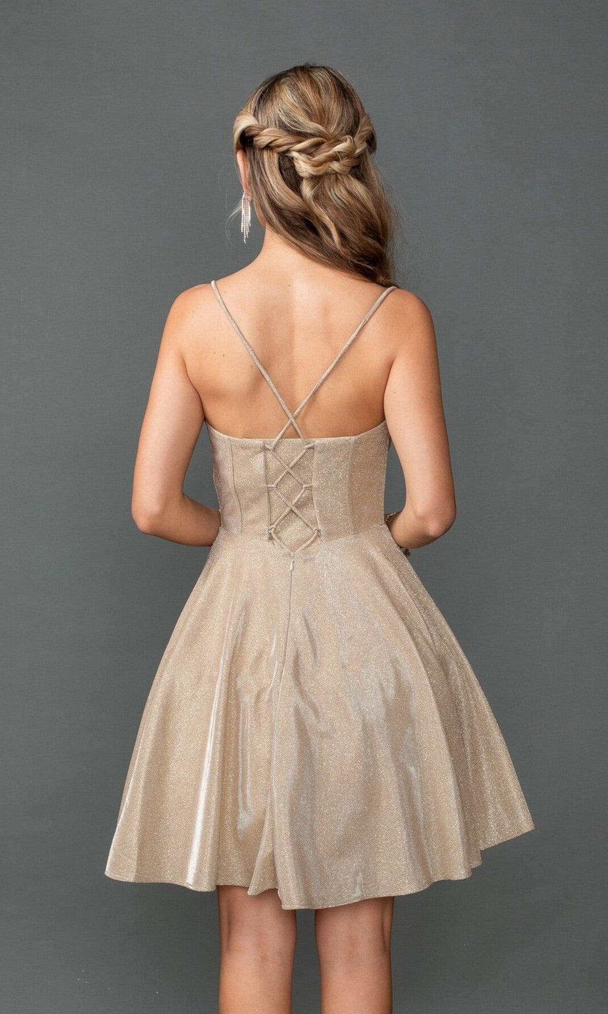Woman modeling the Dancing Queen 3421 short prom dress in sparkly beige glitter with crisscross straps, a lace-up back, and corset bodice, stands against a plain gray background facing away from the camera.